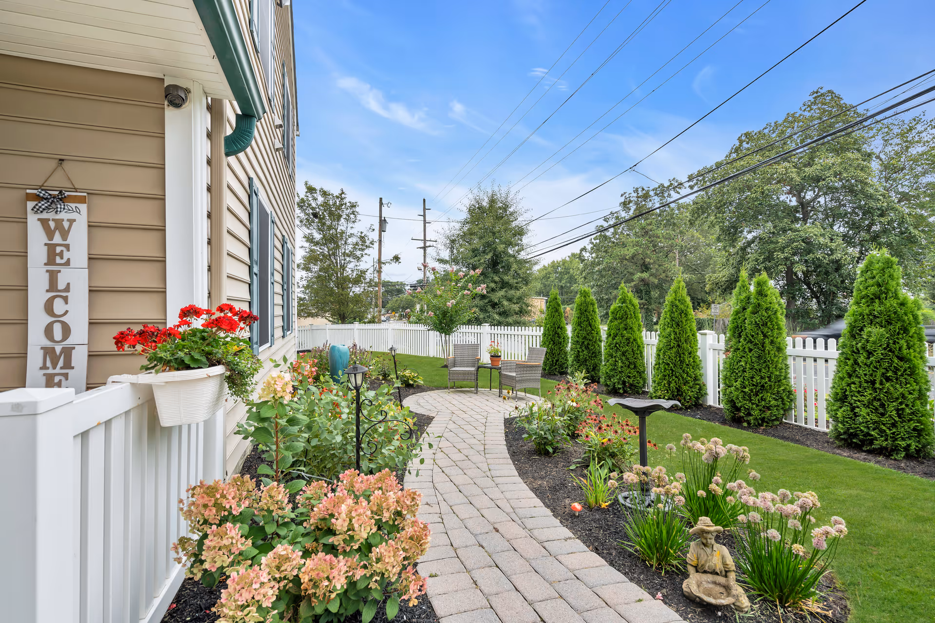 Paved garden walkway beside a beige building with a white picket fence, flowering plants, shrubs and outdoor seating.