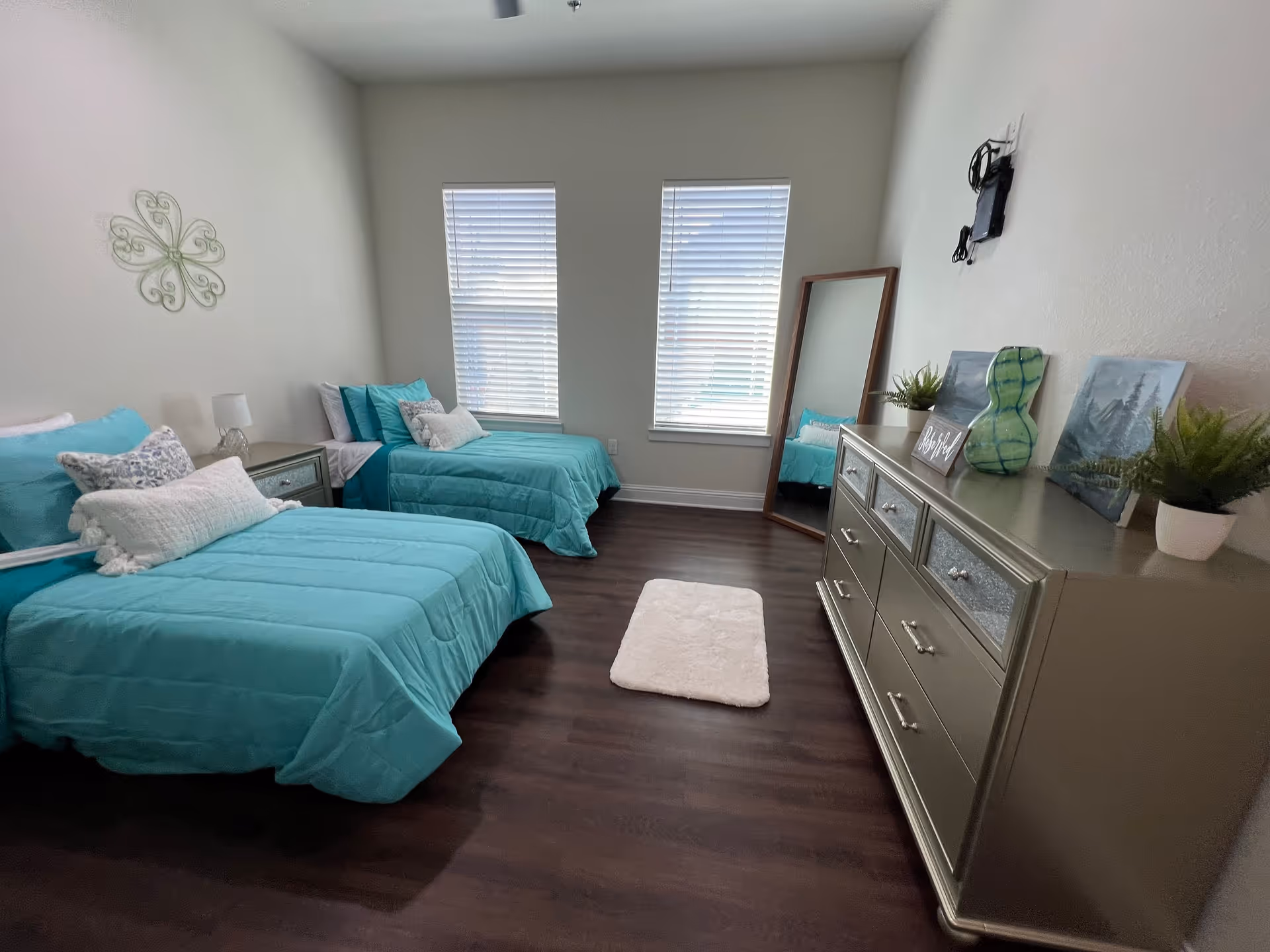 A bedroom with two twin beds dressed in turquoise bedding, each with multiple pillows. Between the beds is a small nightstand with a lamp. On the right side of the room is a large dresser with decorative items including plants, a green glass vase, and artwork. A full-length mirror leans against the wall near two windows with white blinds. The floor is dark wood, and a small white rug is placed in front of the dresser.
