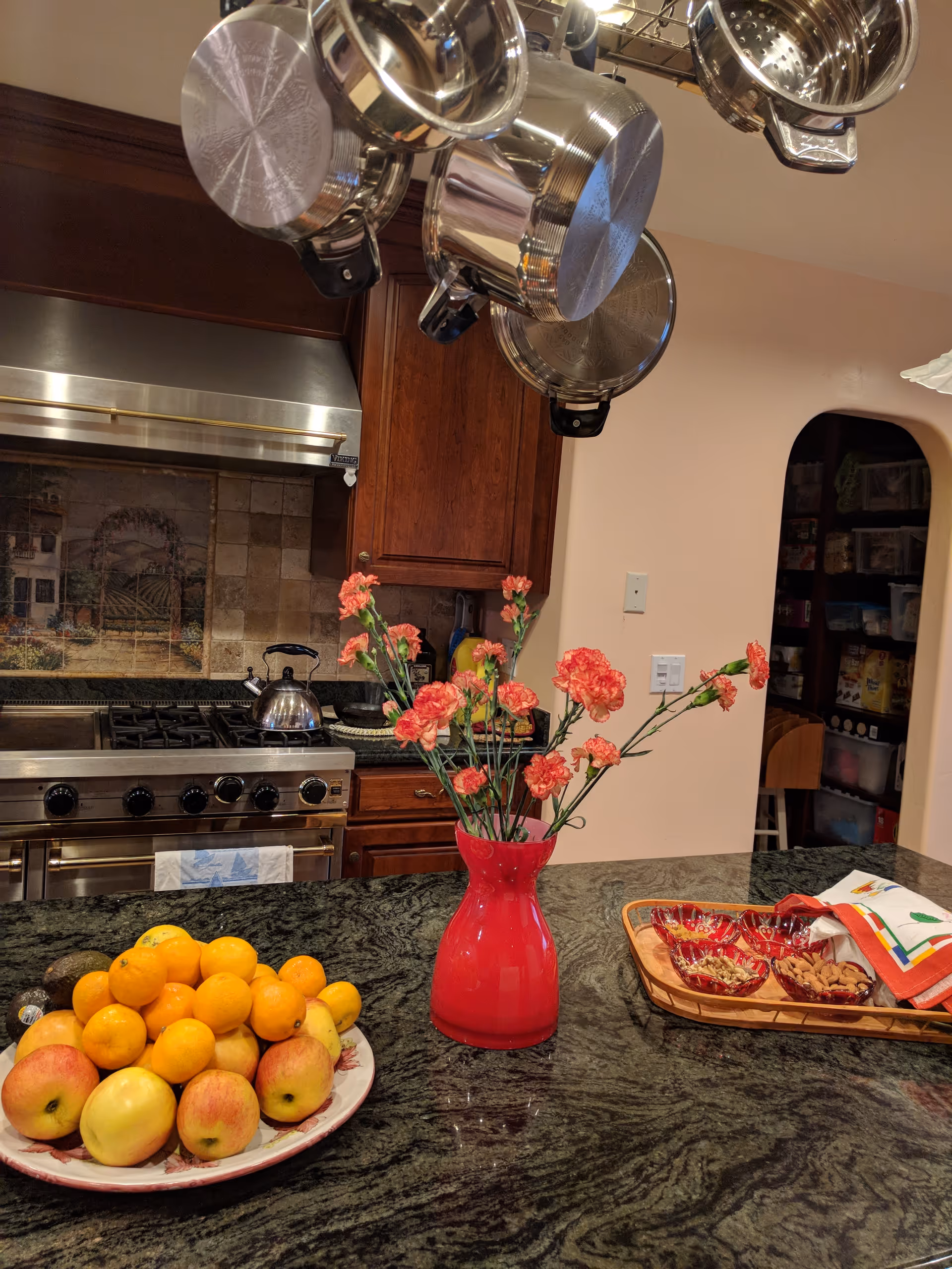 A kitchen interior featuring a dark granite countertop with a red vase holding pink carnations, a plate piled with apples and oranges, and a wooden tray with small bowls of nuts and a folded cloth. Stainless steel pots hang above the counter, and a stove with a kettle is visible in the background along with wooden cabinets and a tiled backsplash.
