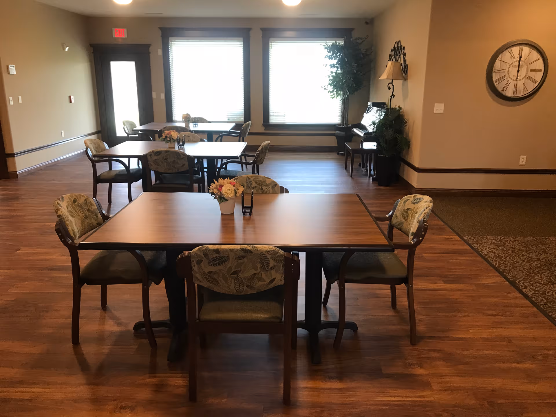 A dining area with wooden tables and cushioned chairs featuring leaf-patterned upholstery. Each table has a small flower arrangement. The room has wooden flooring, large windows with blinds, a piano in the corner, a wall clock, and a door with an exit sign above it.