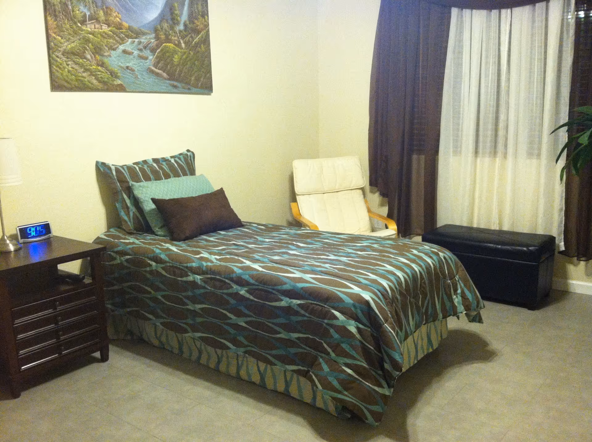 A bedroom with a single bed covered in a brown and blue patterned bedspread, a wooden nightstand with a lamp and digital clock, a white cushioned chair, a black storage bench, brown curtains, and a landscape painting on the wall.