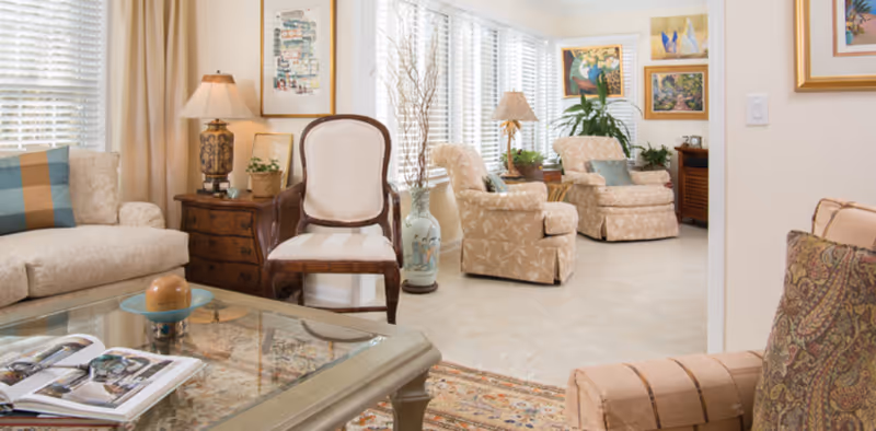 Well-lit living room with upholstered sofas and chairs, a glass coffee table, lamps, plants, and framed artwork.