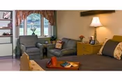 A cozy studio room with a bed, bedside table and lamp, upholstered armchair and loveseat, and a window with a floral valance.
