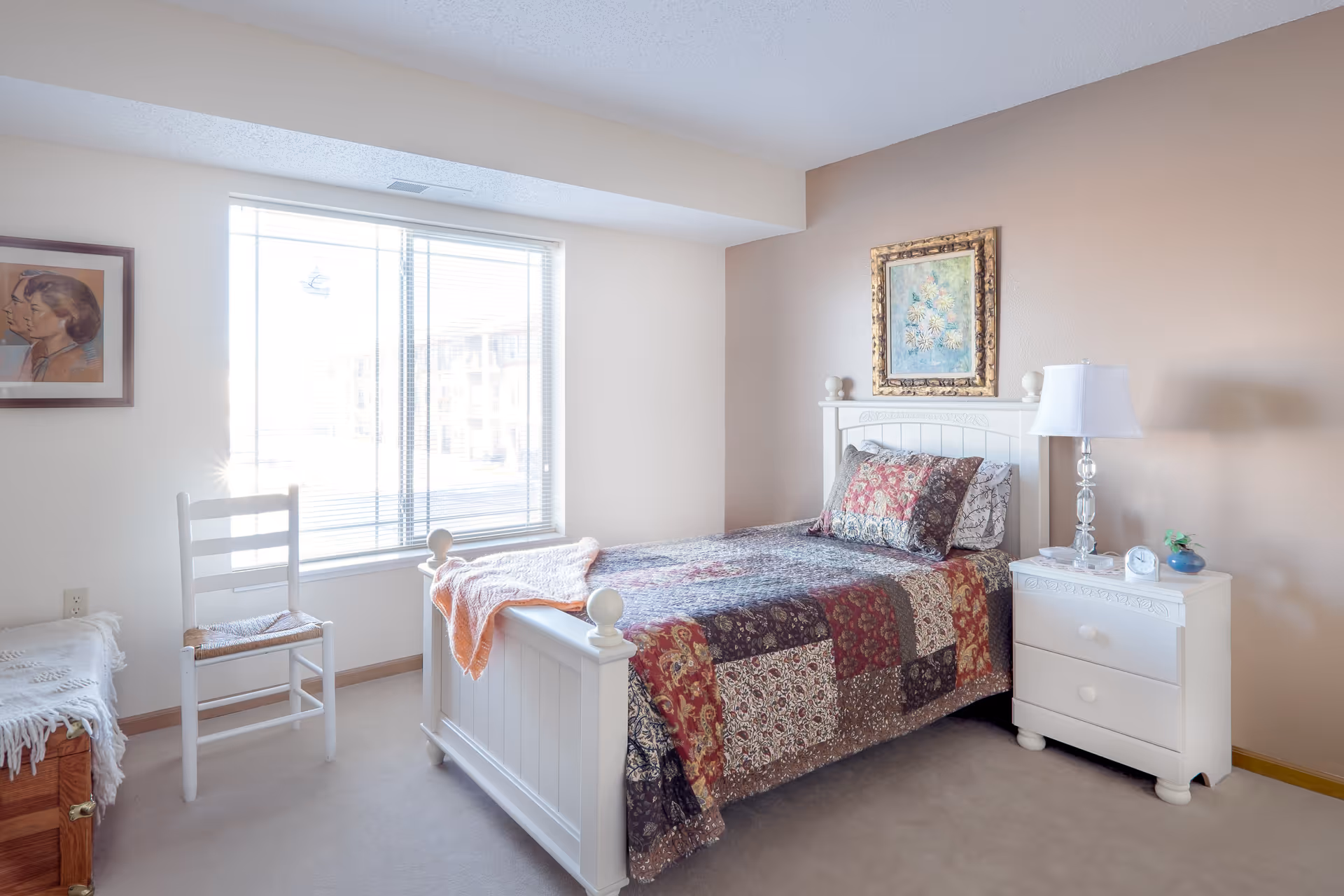 A bright and cozy bedroom with a single white wooden bed featuring a patchwork quilt in red, brown, and beige tones. Next to the bed is a white nightstand with a lamp, clock, and small plant. A white wooden chair is placed near a large window with blinds, and a framed portrait hangs on the wall. The room has beige walls and carpeted floor.