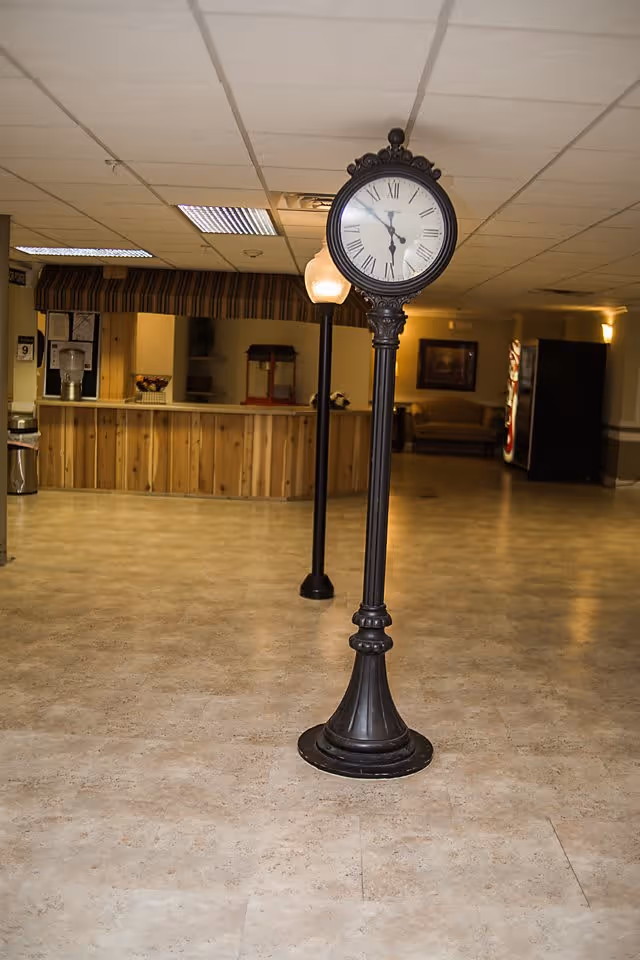 An indoor area with a vintage-style black standing clock showing the time as 11:30. Behind the clock is a wooden counter with a striped valance, a water dispenser, and a popcorn machine. The floor is tiled, and there is a vending machine and a couch in the background under warm lighting.