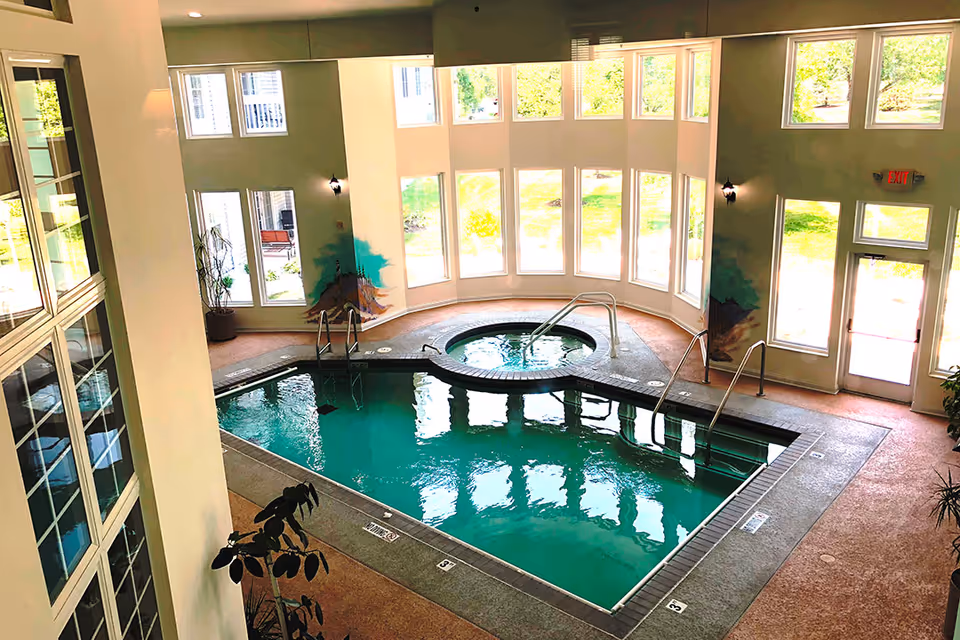 Indoor community pool and circular hot tub in a bright room with large windows and seating area.