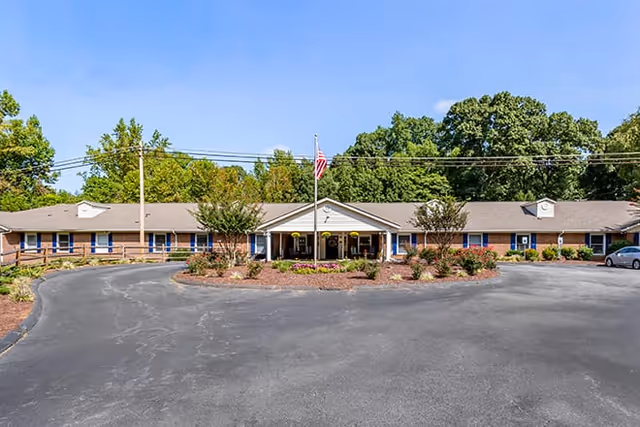 Front exterior view of Brookdale East Broad facility with a circular driveway, landscaped garden with flowers and shrubs, an American flag on a flagpole, and a single-story building with a gray roof and multiple windows under a clear blue sky.