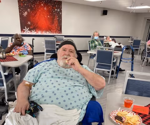 Residents sitting at tables in a communal dining room of a care facility, with snacks on a nearby table.