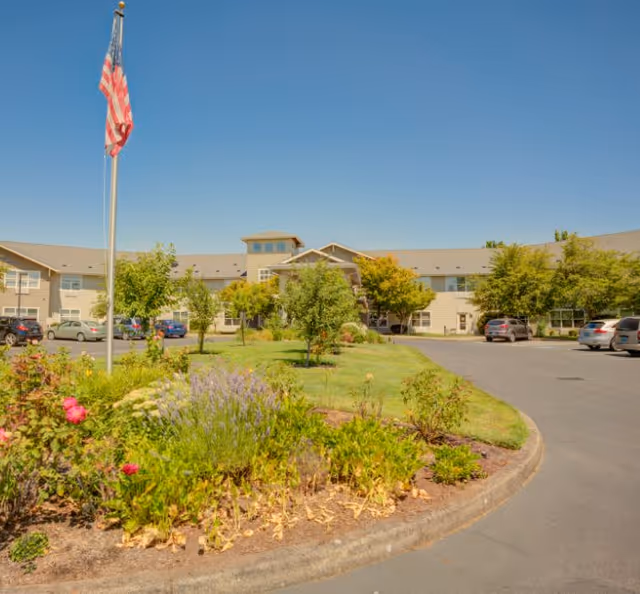 Exterior view of Evergreen Senior Living Eugene facility with a garden featuring various plants and flowers, an American flag on a flagpole, a driveway with parked cars, and a two-story building under a clear blue sky.