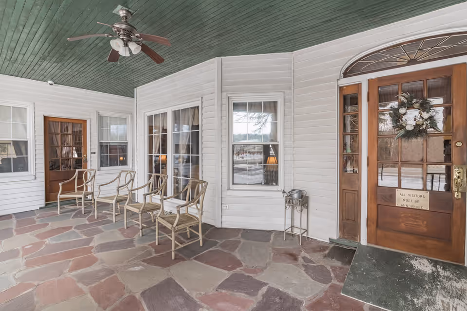 Covered front porch with stone floor, a row of chairs, windows and a wooden entry door decorated with a wreath.