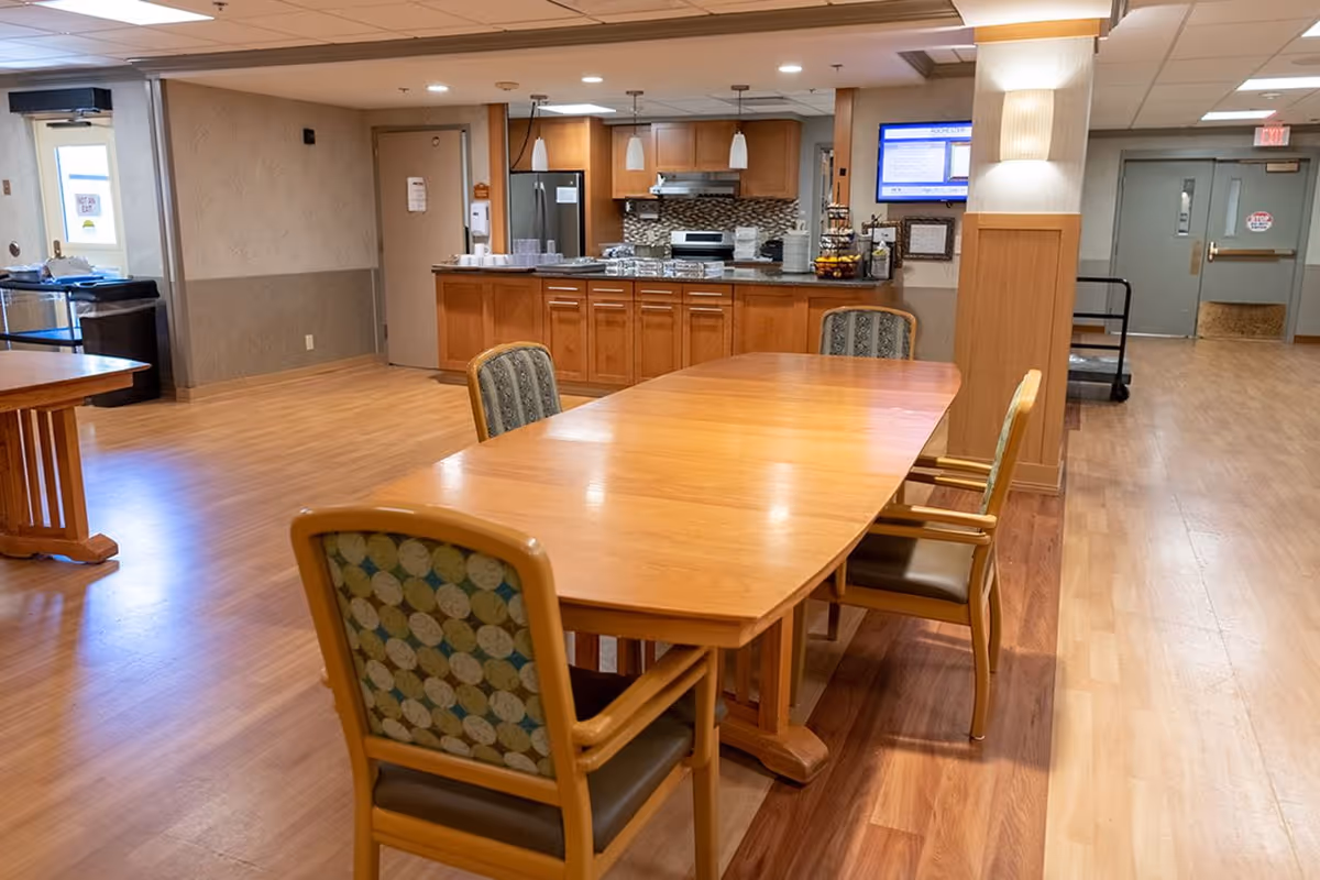 A dining area in a senior living facility featuring a wooden table with four chairs around it. In the background, there is a kitchen area with wooden cabinets, a refrigerator, stove, and countertop with various items. The room has wood flooring and neutral-colored walls, with a hallway and doors visible in the background.