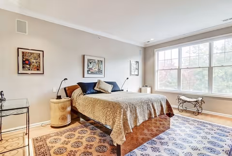 Sunlit bedroom featuring a bed with patterned bedding, framed artwork, area rugs, side tables, and a large window.