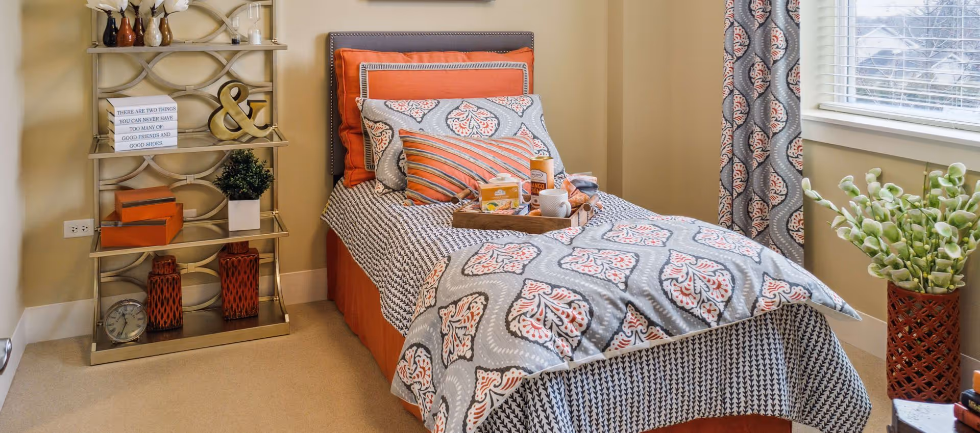 A neatly made single bed with patterned bedding and multiple pillows in shades of orange, gray, and white. A wooden tray with snacks and a cup is placed on the bed. To the left of the bed is a decorative shelving unit with various items including a clock, vases, and a sign that reads 'There are two things you can never have too many of: good friends and good shoes.' To the right is a window with patterned curtains and a tall vase with green plants.