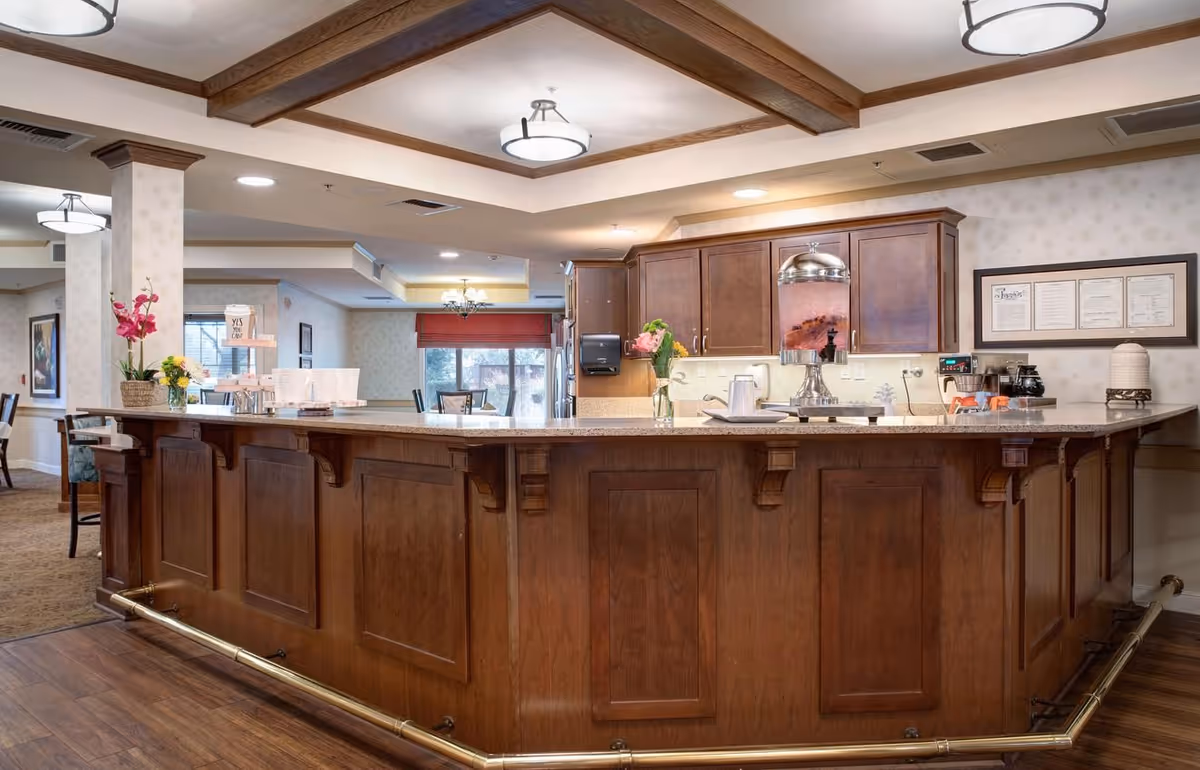 Wood-paneled service counter and kitchenette area with pendant lights and seating in a senior living dining/lounge space.