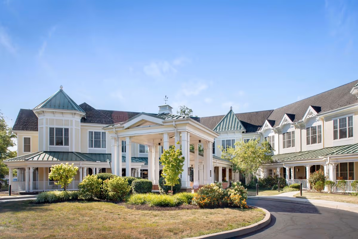 Exterior view of a large, two-story senior living facility building with white columns, green metal roofs, and multiple windows. The entrance features a covered driveway with a circular driveway and landscaped bushes and trees under a clear blue sky.