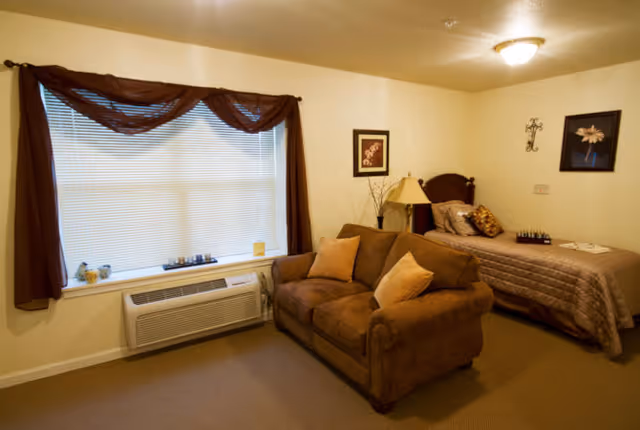 A cozy room with a single bed covered in a brown quilt, a brown loveseat with two beige pillows, a window with closed blinds and brown curtains, a wall lamp, framed pictures, and a beige carpeted floor.