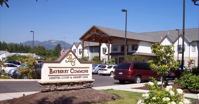 Exterior view of Bayberry Commons assisted living and memory care facility with a large sign in the foreground, parked cars, and a two-story building with a covered entrance under a clear blue sky.