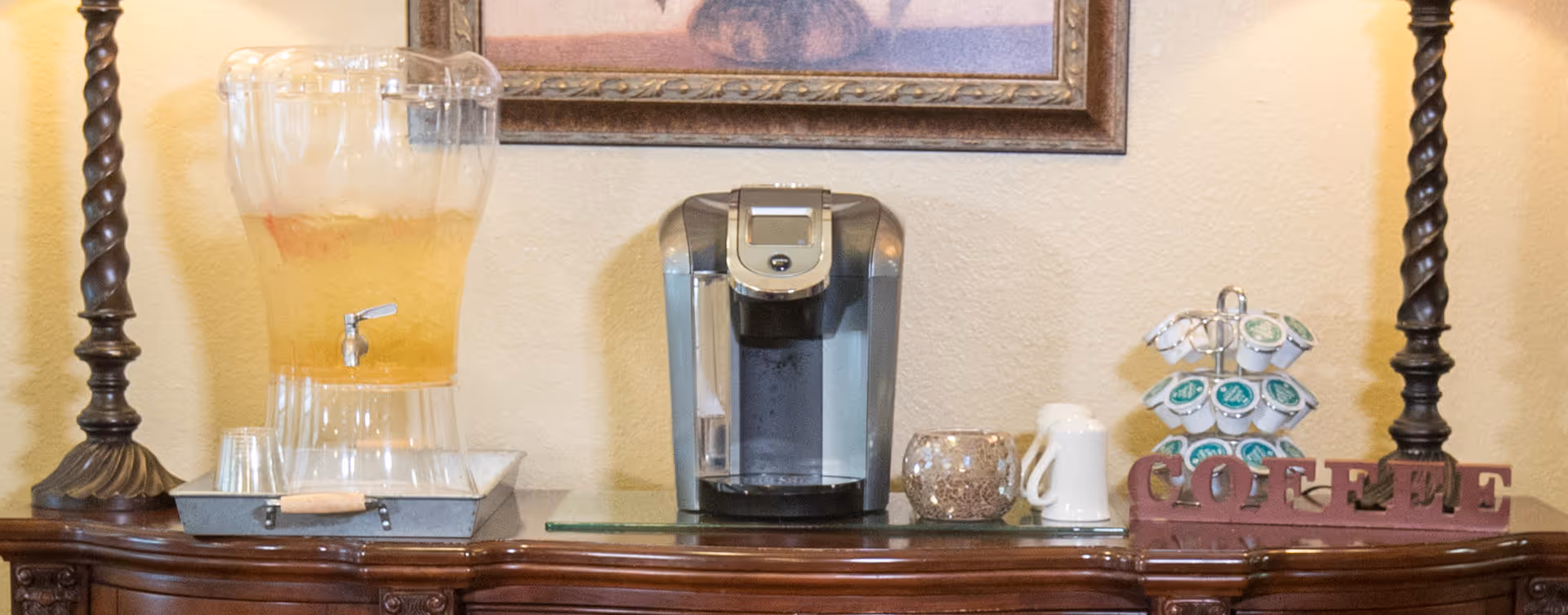 A coffee and beverage station on a wooden sideboard with a glass container of iced tea with a spigot, a coffee maker, a decorative candle holder, a white ceramic pitcher, and a rotating stand holding coffee pods. The word 'COFFEE' is displayed in large letters on the right side of the sideboard, with two tall twisted wooden lamps on either end and a framed picture hanging on the wall above.