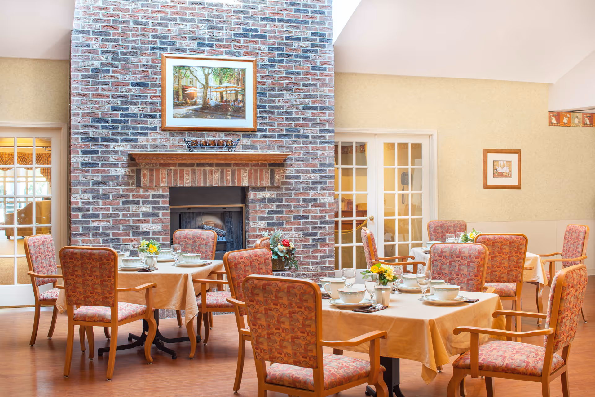 A cozy dining room with several tables covered in beige tablecloths, each set with bowls, glasses, and silverware. The chairs have wooden frames and patterned upholstery. A large brick fireplace with a framed painting above it is centered on one wall. The room has warm lighting and glass-paneled doors leading to other areas.