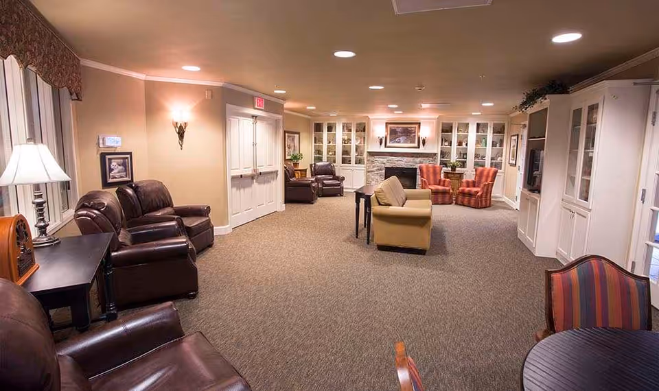 A spacious, well-lit living room area in a senior living facility with various seating options including leather armchairs, a beige sofa, and striped upholstered chairs. The room features built-in white cabinetry, a stone fireplace with a framed picture above it, and soft wall lighting. There is a round table with chairs in the foreground and large windows with curtains on the left side.