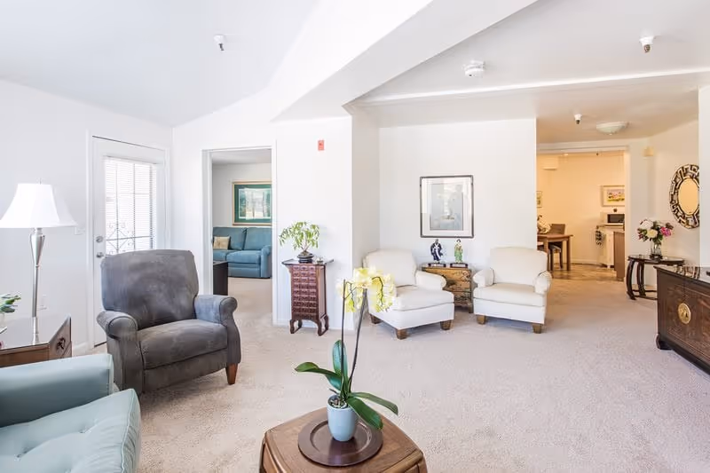 A bright and spacious living room area with beige carpet and white walls. The room features a gray armchair, two white armchairs, a wooden coffee table with a potted orchid, and various decorative items including a lamp, framed artwork, and a mirror. In the background, there is a view into an adjacent room with a blue sofa and a dining area with a table and chairs.