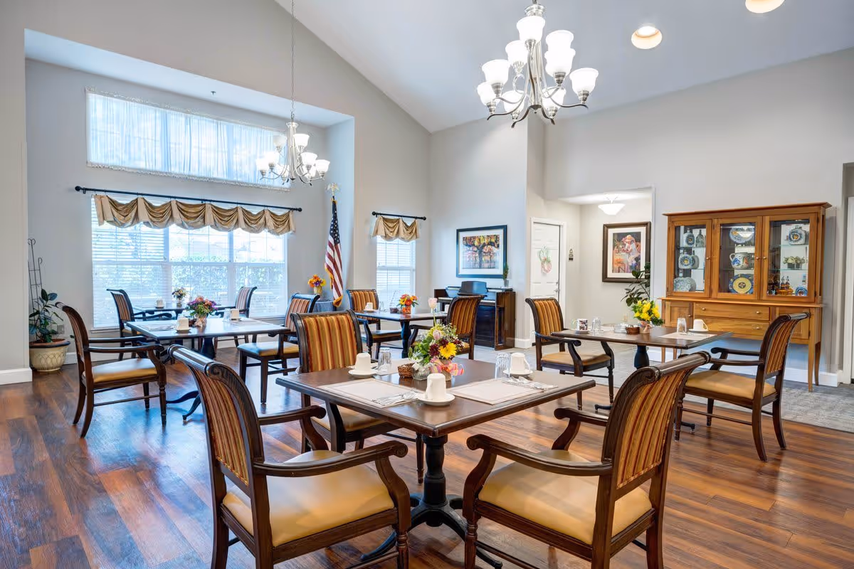 A bright dining room with several wooden tables and chairs arranged neatly. Each table is set with white cups, glasses, and placemats, and decorated with small flower arrangements. Large windows with beige valances allow natural light to fill the room. The room features hardwood floors, a chandelier, framed artwork on the walls, and a wooden cabinet displaying decorative plates and items. An American flag stands near the windows.