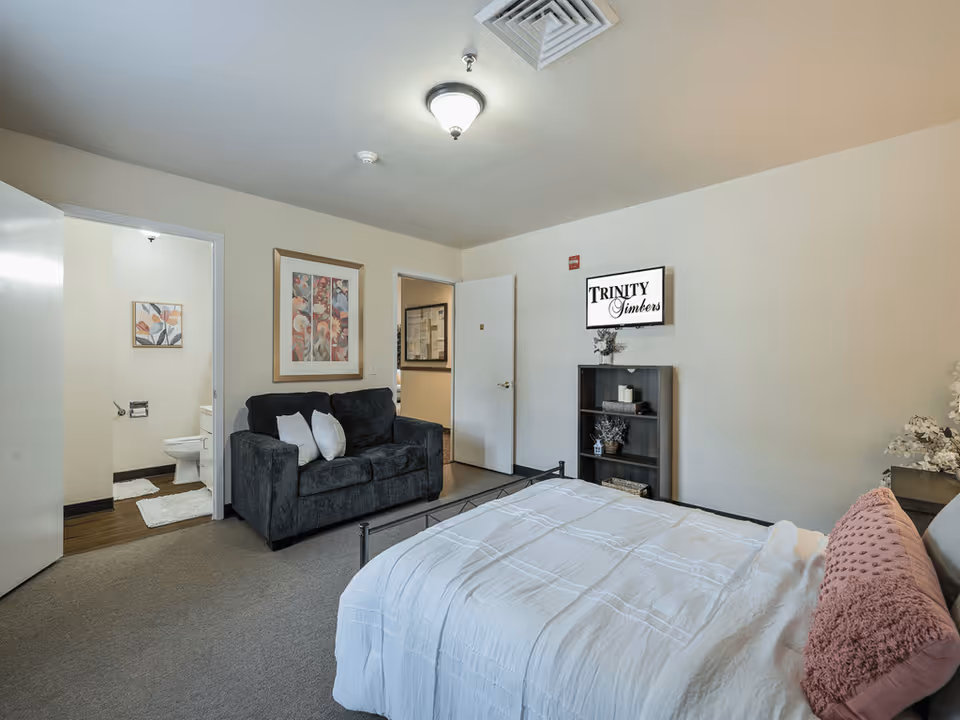 A furnished assisted-living bedroom with a bed, sofa, shelving unit, open bathroom doorway, and a wall sign that says 'Trinity Timbers'.