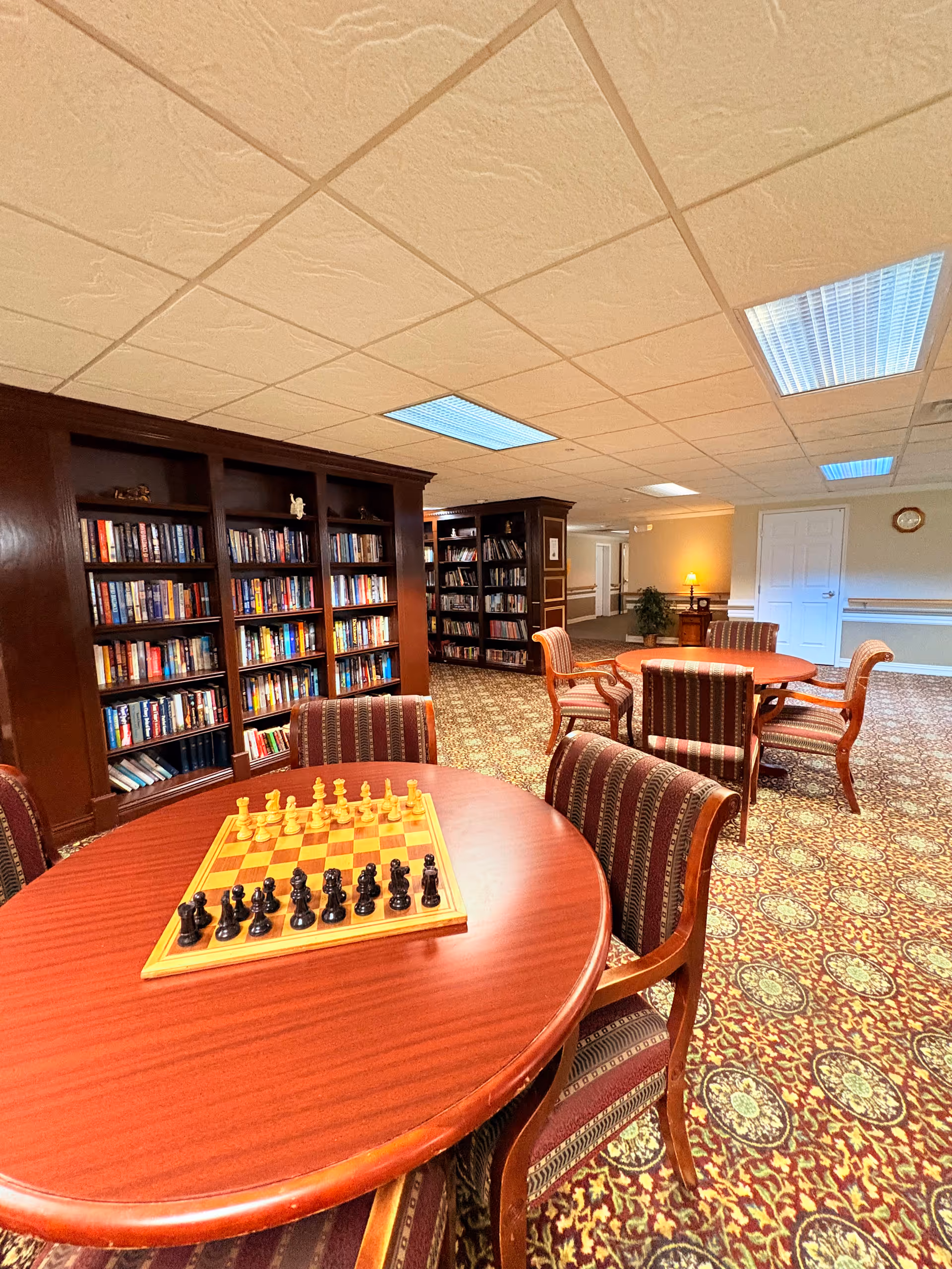 A cozy common area in an assisted living facility featuring round wooden tables with striped upholstered chairs. One table has a chessboard set up for a game. In the background, there are large wooden bookshelves filled with books. The room has patterned carpet, a drop ceiling with fluorescent lights, and a door on the far wall.