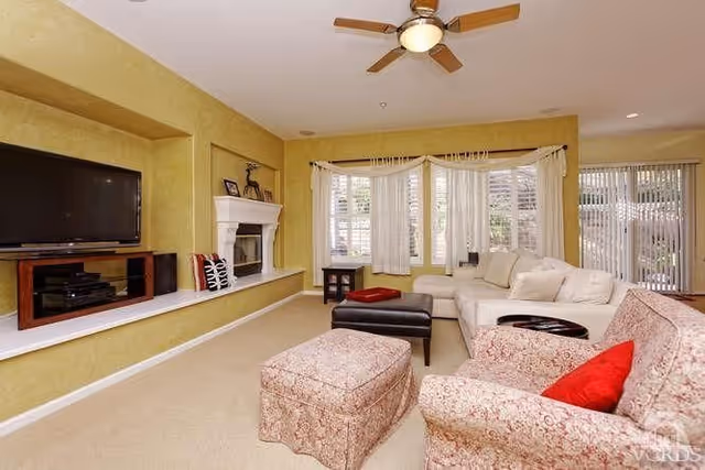 Bright living room with a sectional sofa, patterned armchair and ottoman, fireplace, wall-mounted TV, and ceiling fan.