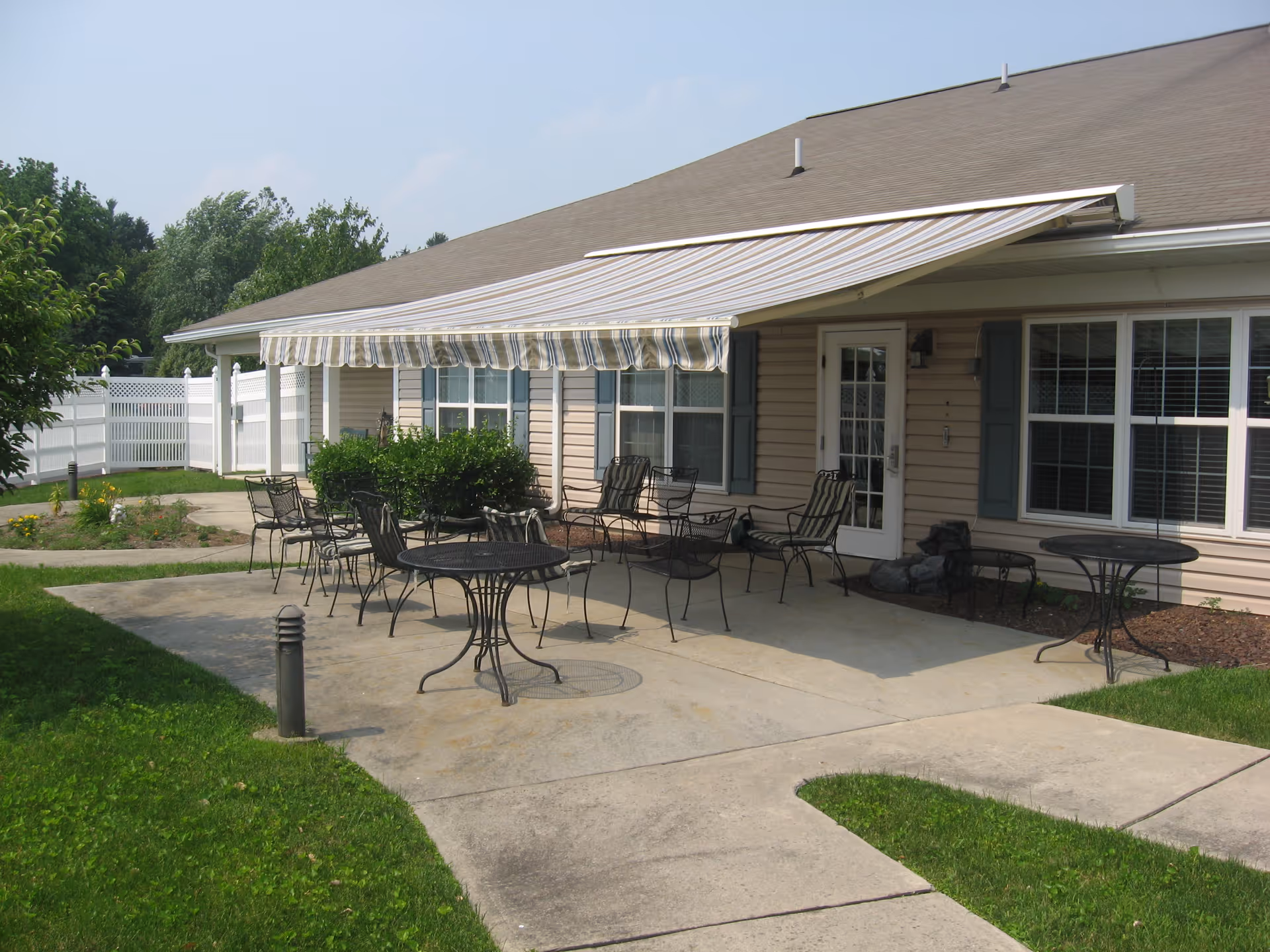 Outdoor patio area at Linden Village A ProMedica Senior Living Community with metal tables and chairs on a concrete surface, a retractable striped awning providing shade, beige siding building with windows and a door, surrounded by green grass and a white fence.
