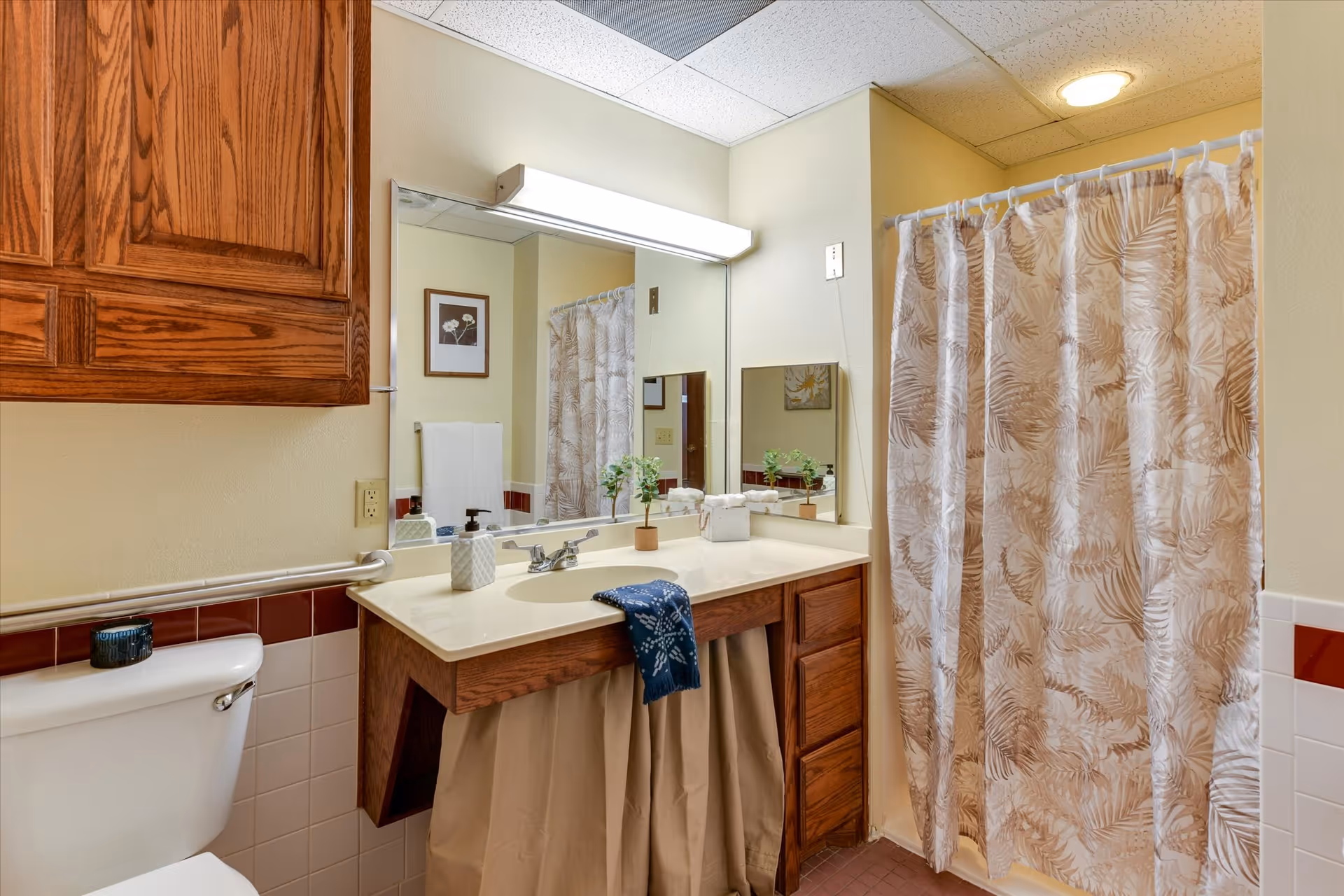Bathroom with a white toilet, wooden cabinets, a sink with a beige countertop, a large mirror, and a shower with a beige and white patterned curtain. The walls are painted light yellow and there is a framed picture and a towel hanging on the wall.