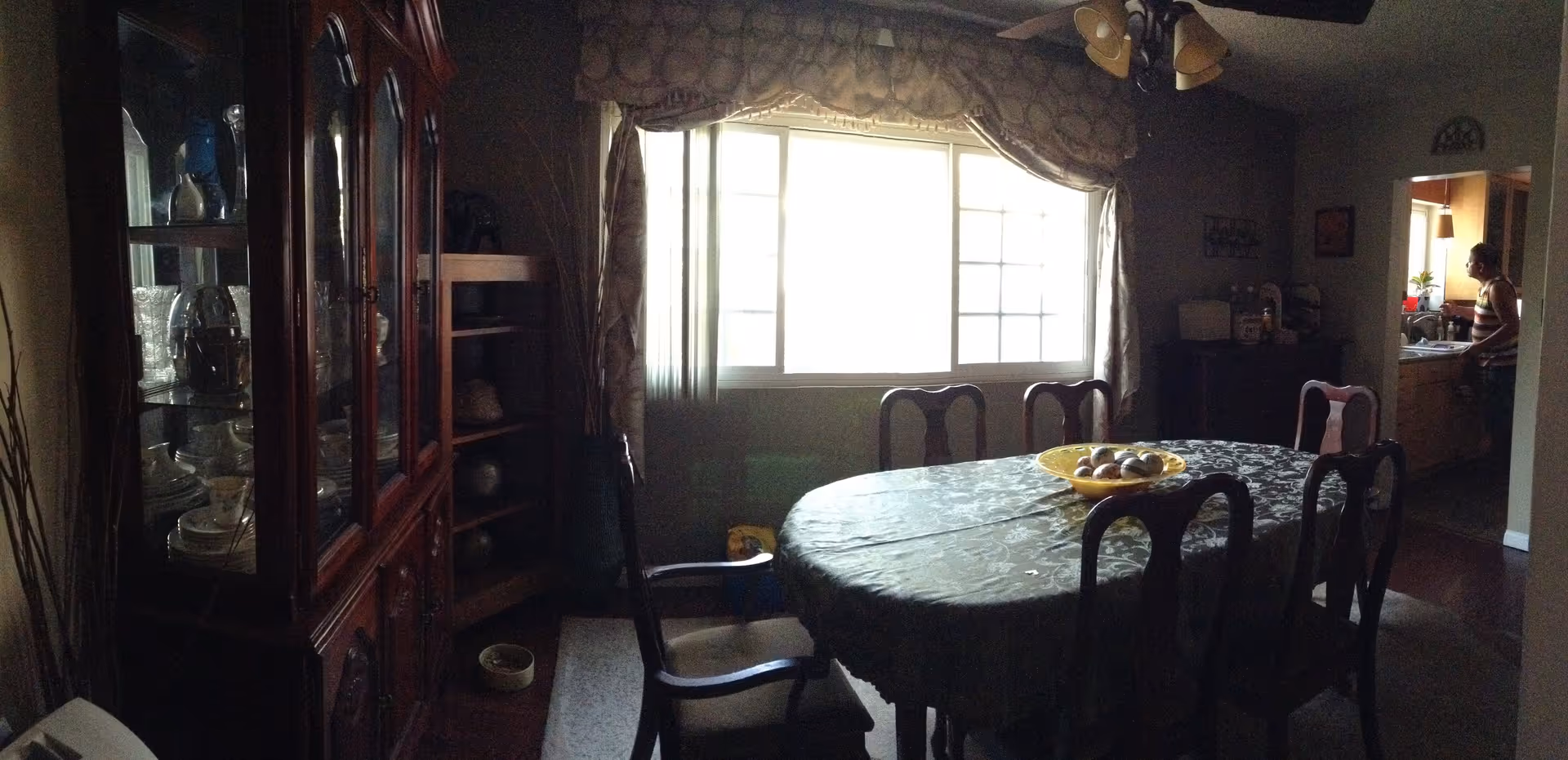 A dining room with a large oval table covered with a patterned tablecloth and surrounded by six wooden chairs. A yellow bowl with decorative balls is placed in the center of the table. To the left, there is a wooden china cabinet displaying glassware and dishes. A large window with curtains allows natural light into the room. In the background, a person is visible in the adjacent kitchen area.