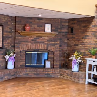 Cozy interior corner featuring a brick fireplace with a wooden mantel. On the mantel, there is a red decorative vase and a framed picture. On either side of the fireplace, there are built-in brick seating areas with flower arrangements in white vases. To the right, a white side table holds a small potted plant and decorative items. The floor is wooden, and the ceiling has recessed lighting.