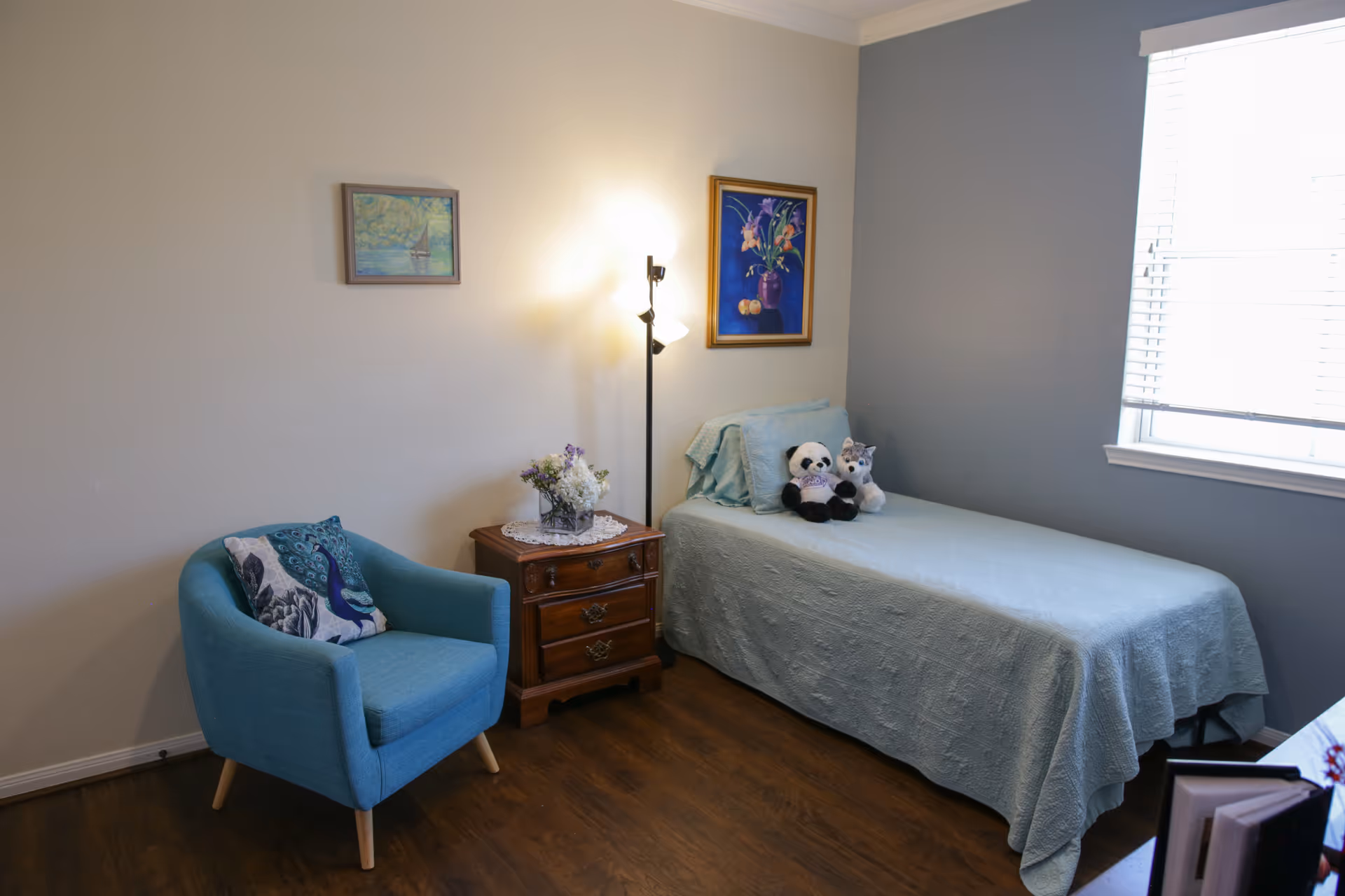 A tidy single bedroom with a twin bed topped with stuffed animals, a teal armchair, a wooden nightstand with a lamp and flowers, and a window with blinds.
