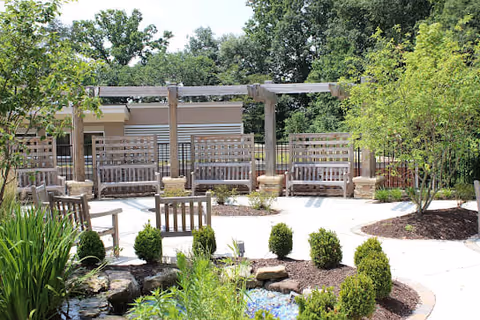 Outdoor seating area with wooden benches arranged in a semi-circle under a wooden pergola. The area is surrounded by greenery including small bushes, trees, and a small pond with rocks. The setting is peaceful and shaded with a paved walkway.