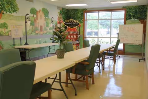 A bright room with a long rectangular table surrounded by green cushioned chairs. There is a potted plant on the table. The walls feature a mural with trees and a building, and there are two whiteboards on stands. Large windows let in natural light and show greenery outside.