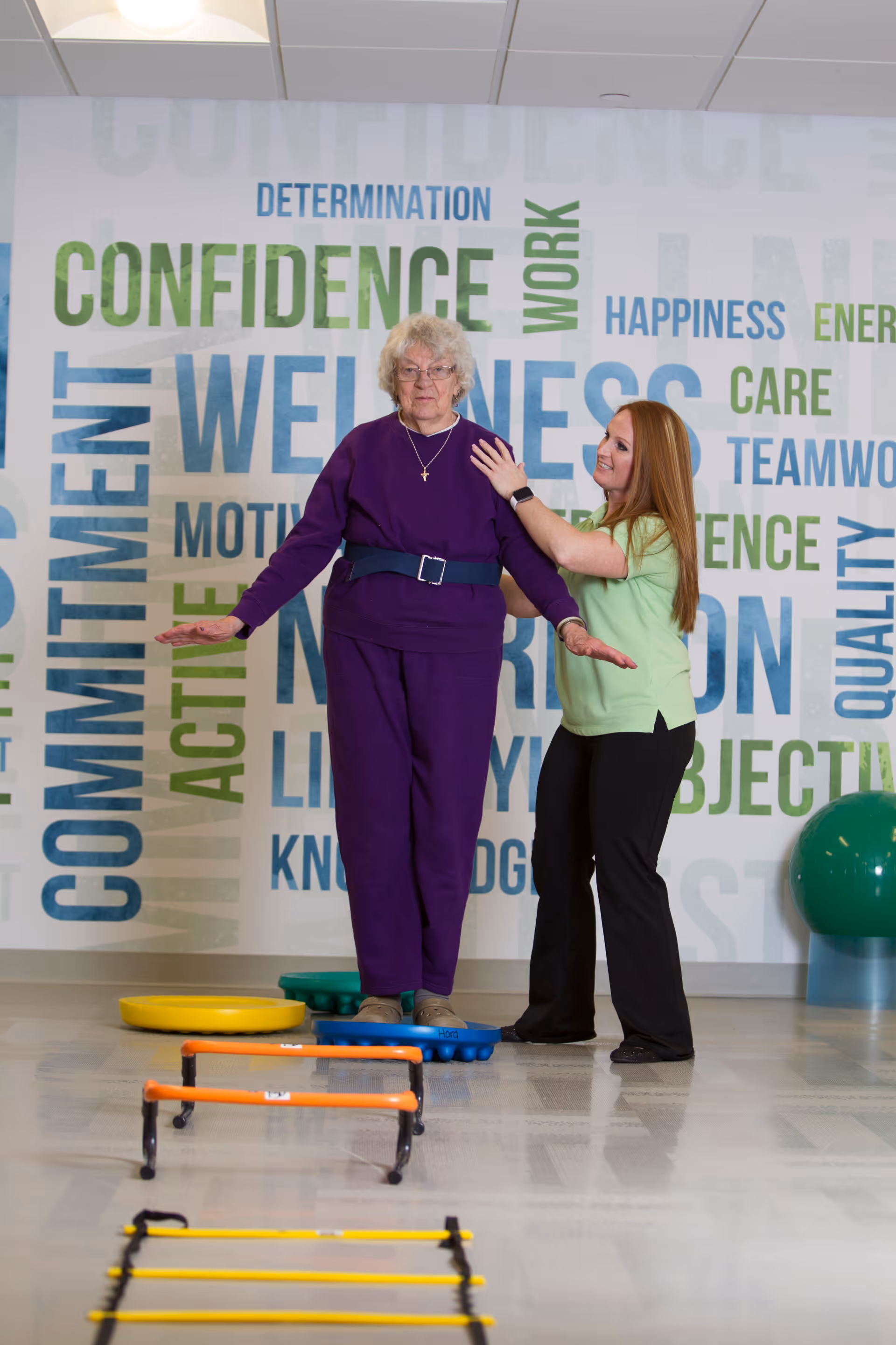 An elderly woman in a purple outfit is balancing on a blue balance pad while being assisted by a caregiver wearing a light green shirt and black pants. They are in a wellness or rehabilitation room with motivational words like 'confidence,' 'wellness,' and 'commitment' on the wall behind them. Exercise equipment such as agility ladders and hurdles are visible on the floor.