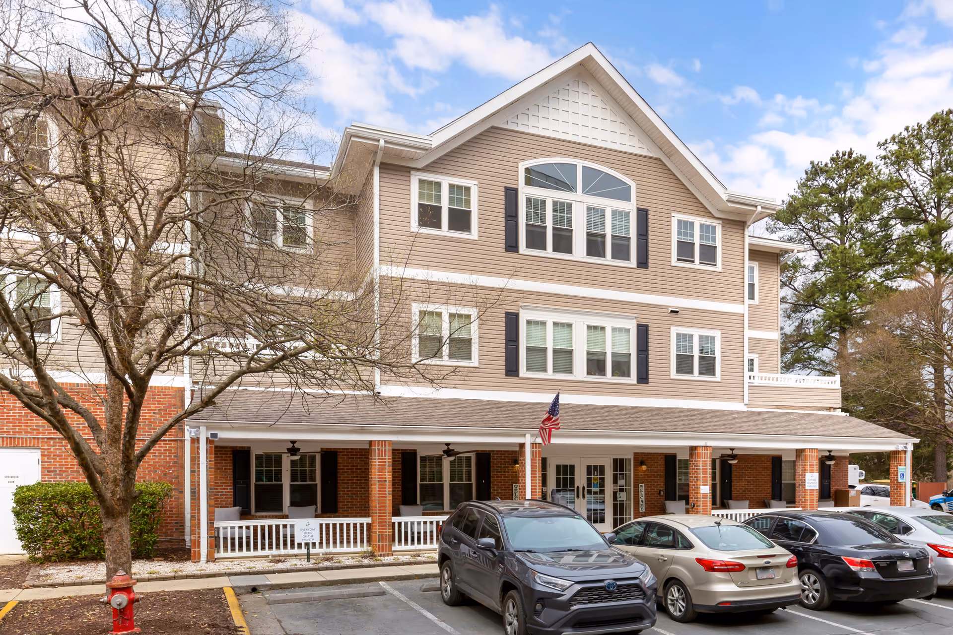 Front exterior of a three-story senior living building with a covered entrance, parked cars, and an American flag.