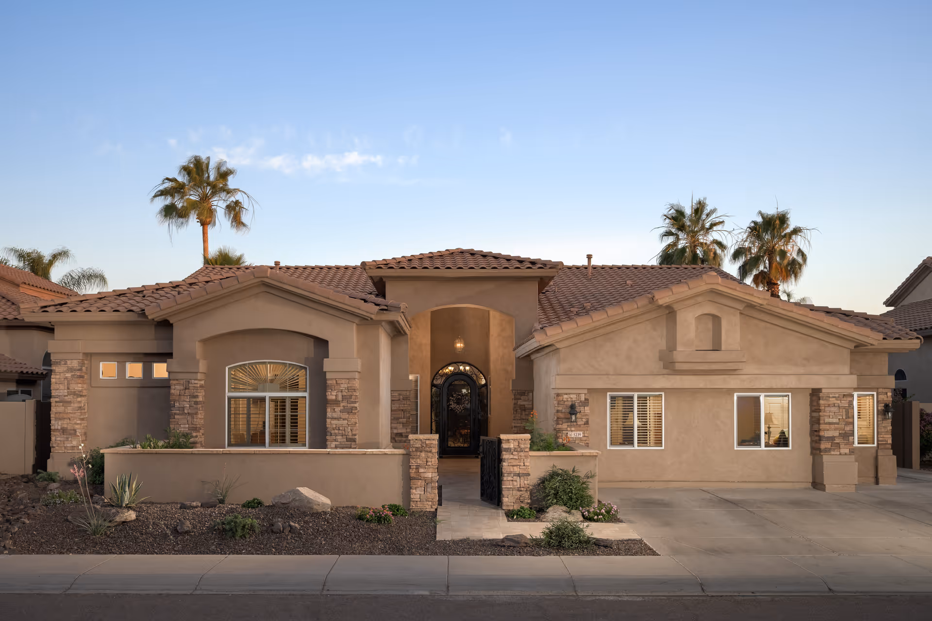 Front exterior view of a single-story house with a tiled roof, stone accents, and a gated entrance pathway. Palm trees are visible in the background under a clear sky.