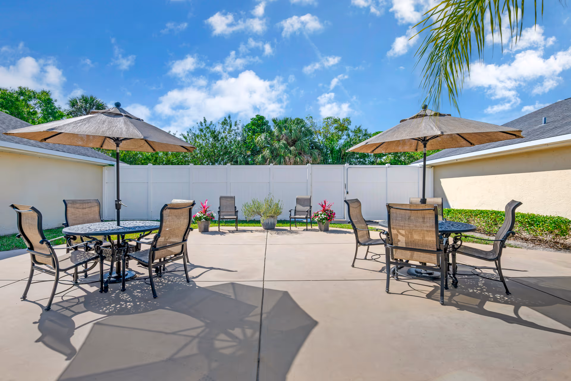 Sunny outdoor patio with round tables, umbrellas, and chairs on a concrete courtyard with potted plants and a white fence.
