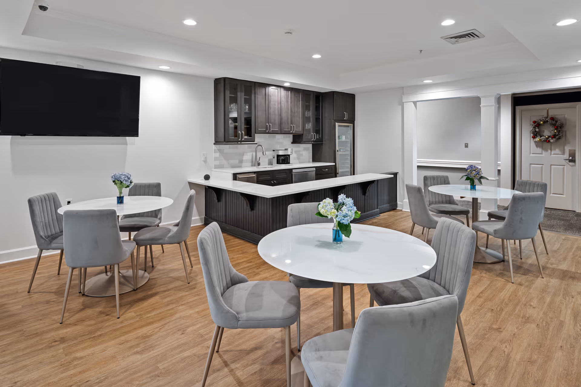 A modern dining area with round white tables and gray upholstered chairs. Each table has a small vase with blue and white flowers. In the background, there is a kitchen area with dark wood cabinets, a white countertop, a microwave, and a refrigerator. A large flat-screen TV is mounted on the wall to the left. The floor is wood, and the walls are painted white with recessed lighting in the ceiling.