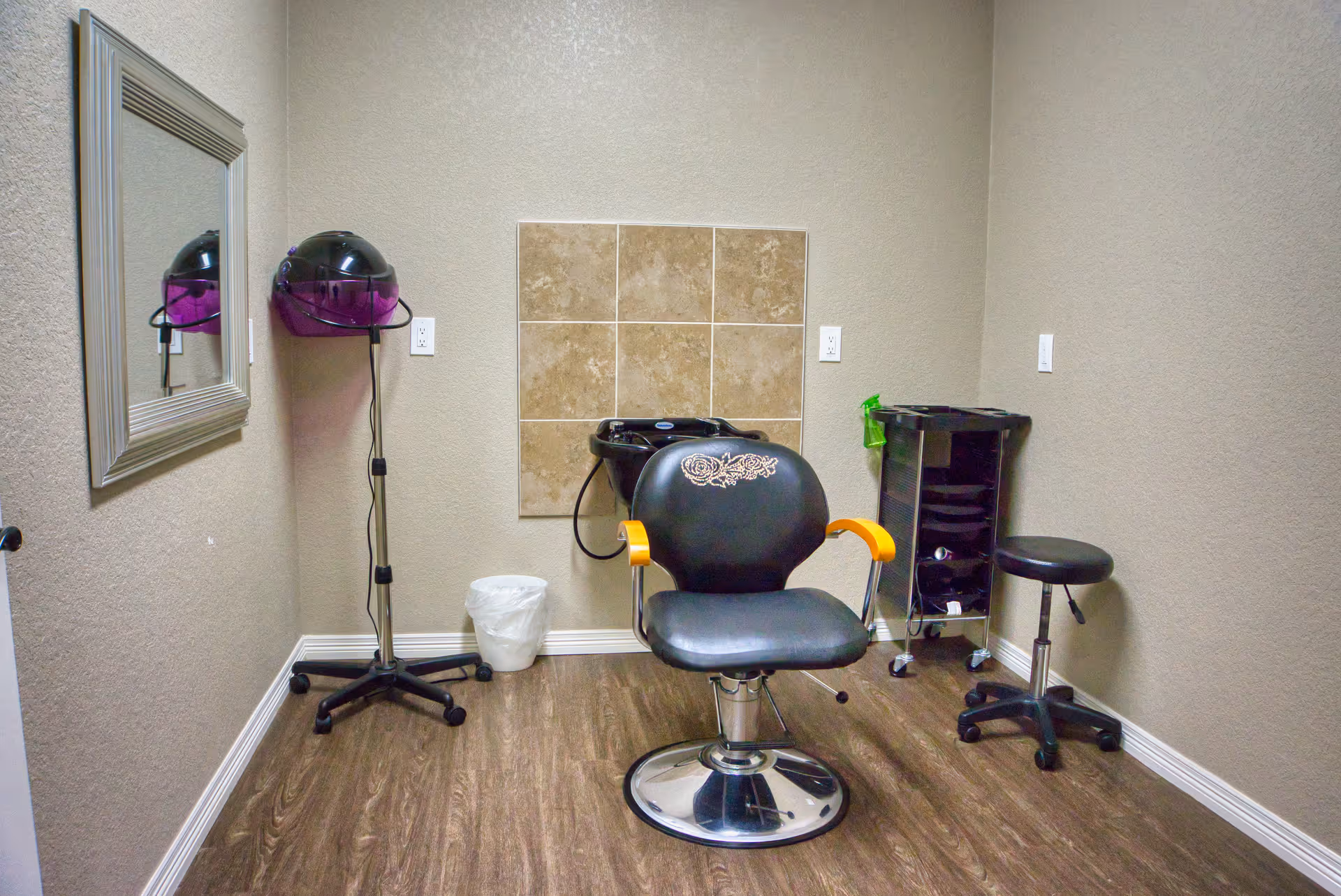 A small hair salon room with a black salon chair featuring orange armrests, a black hair washing sink behind the chair, a black rolling stool, a black rolling cart with hair supplies, a wall-mounted hair dryer with a purple hood, a large mirror on the left wall, beige walls, and wood flooring.