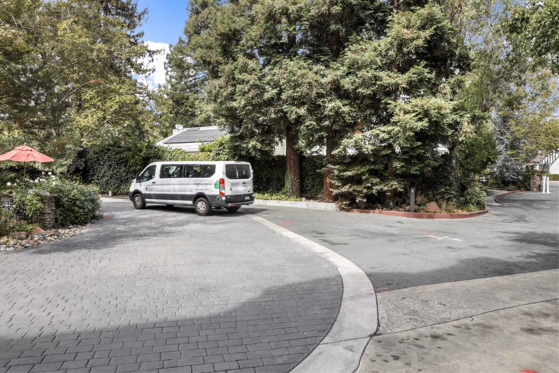 A white passenger van driving on a paved driveway surrounded by tall trees and greenery. There is a red umbrella and some outdoor seating on the left side, and a house roof with solar panels is visible in the background under a partly cloudy sky.