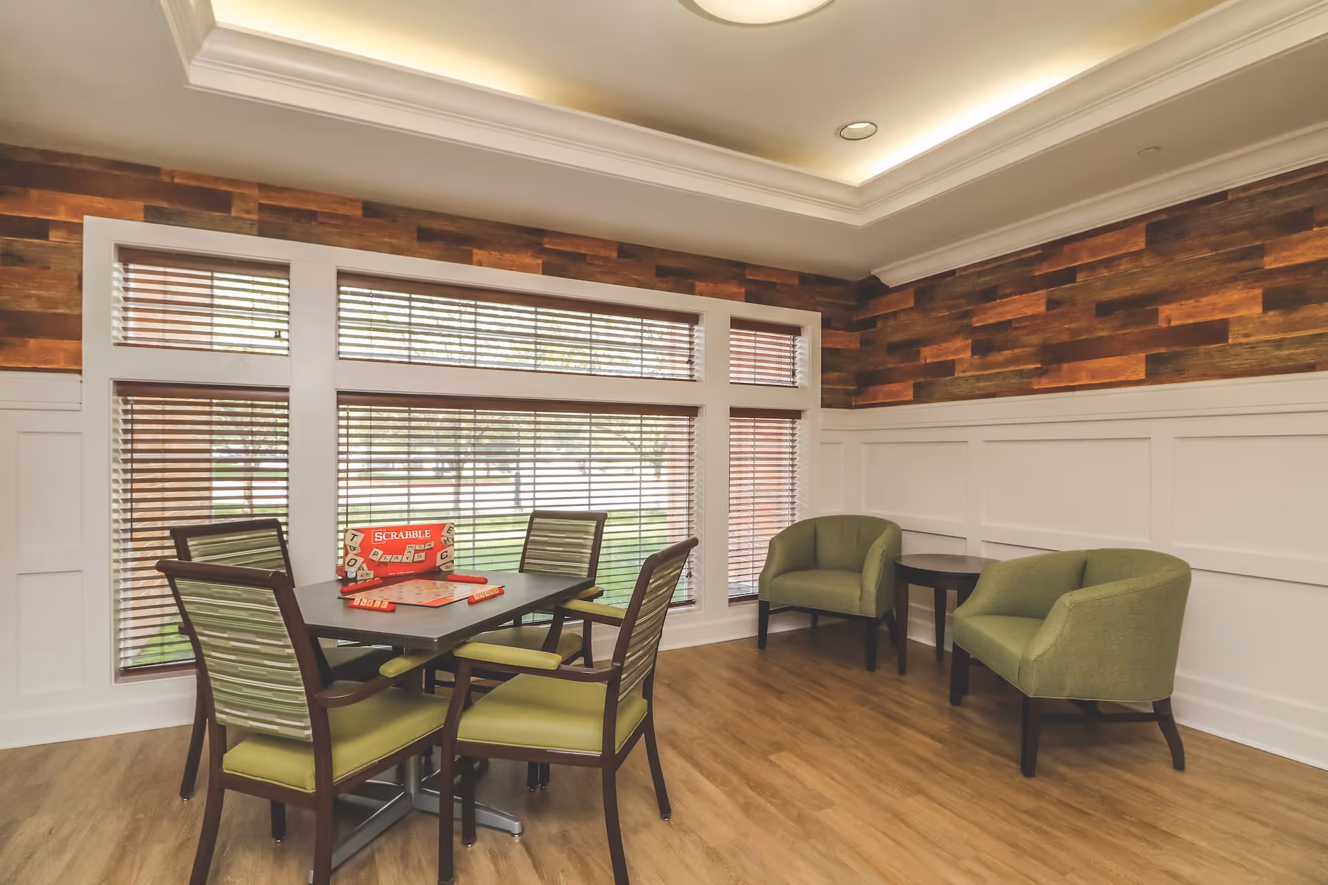 A cozy common area with a table and four chairs arranged for playing Scrabble, with the game set up on the table. The room features large windows with wooden blinds, wood-paneled walls, and two green armchairs with a small side table in the corner. The floor is wooden, and the ceiling has recessed lighting.