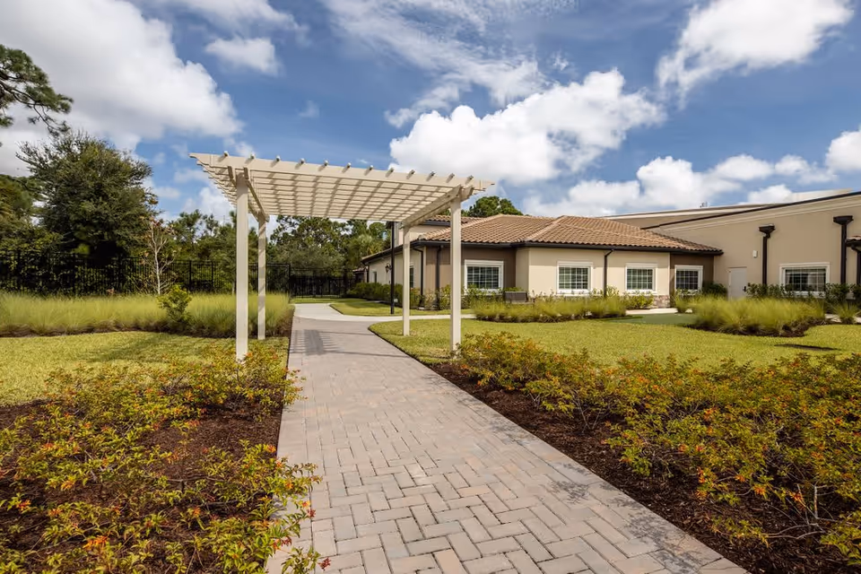 A paved walkway leading through a garden area with low shrubs and grass, featuring a white pergola structure. In the background, there is a single-story building with beige walls and a brown tiled roof under a partly cloudy blue sky.