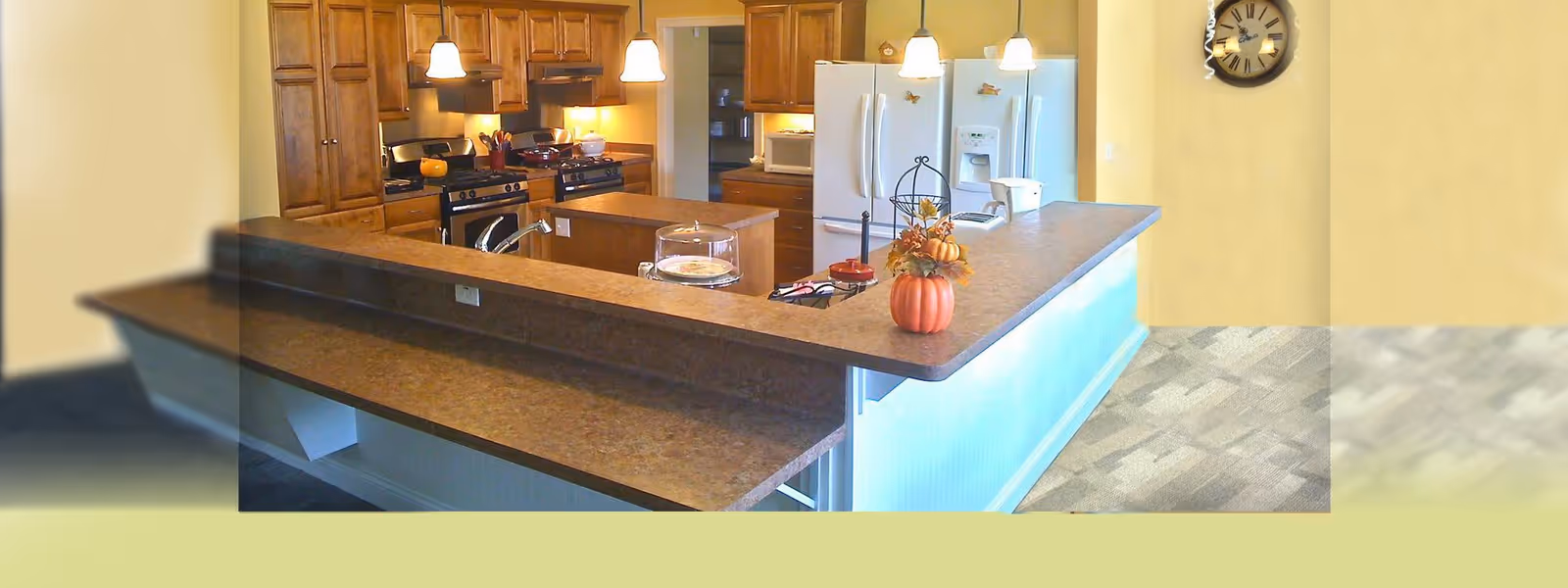 A spacious kitchen with wooden cabinets, two white refrigerators, a stove, and a large countertop with a sink. The countertop has a small pumpkin decoration and a glass cake stand. Three pendant lights hang from the ceiling, and a wall clock is visible on the right wall.
