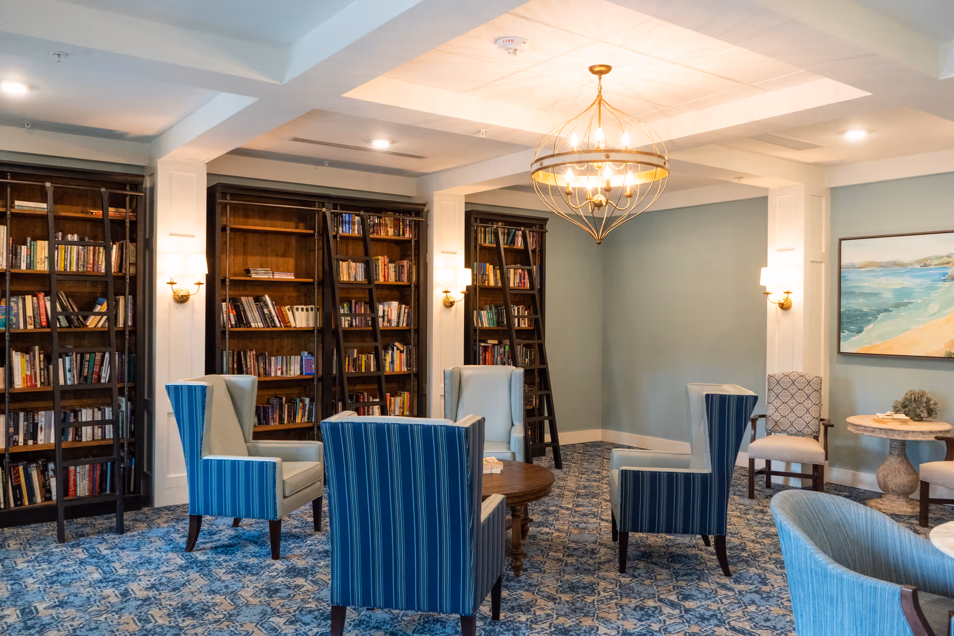 A cozy library room with blue patterned carpet, several blue and beige upholstered armchairs arranged around a wooden coffee table, tall wooden bookshelves filled with books, and a large chandelier hanging from the ceiling. The walls are painted light blue and decorated with a framed painting of a beach scene.
