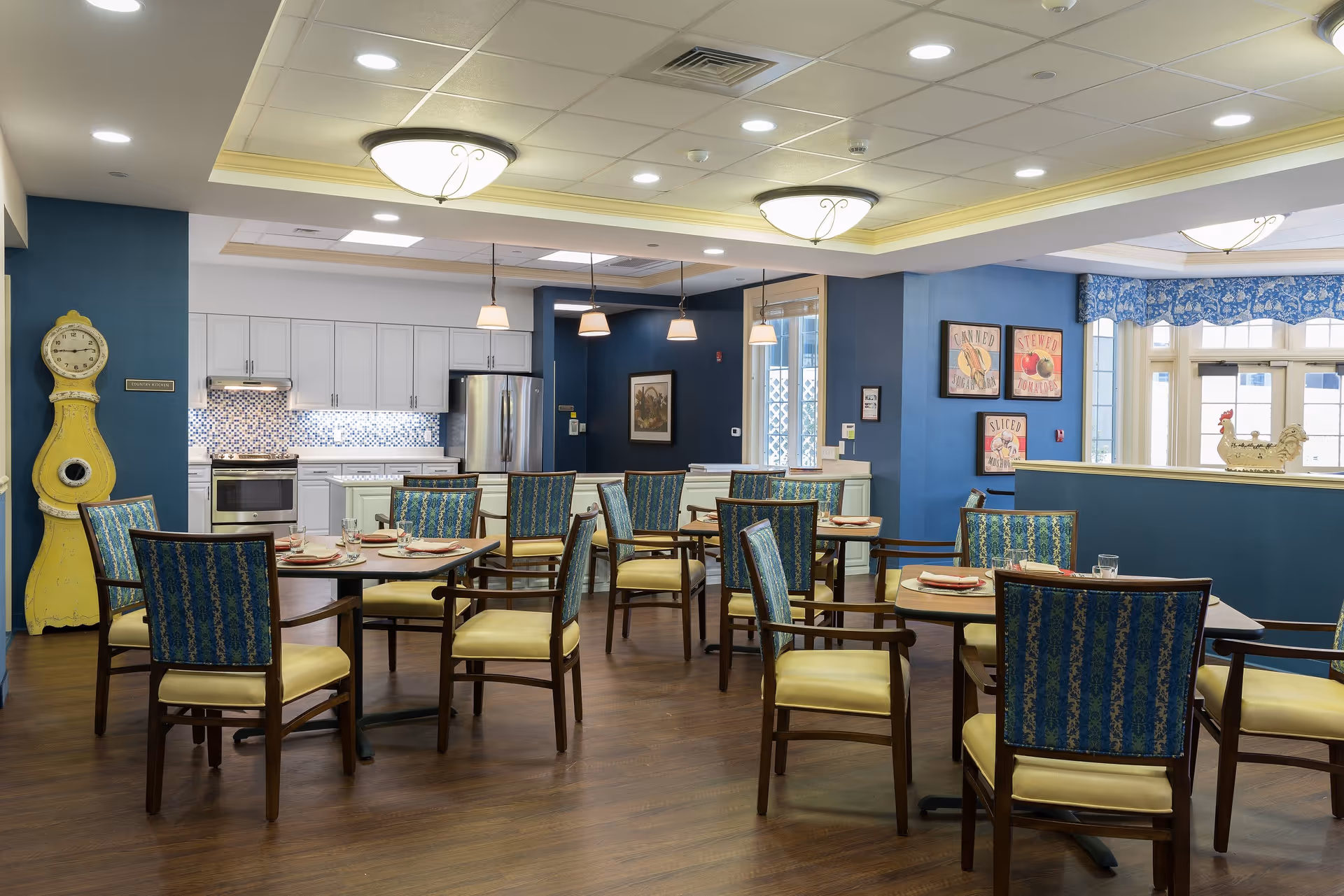 A bright and inviting dining area with several tables and chairs arranged neatly. The chairs have blue patterned backs and yellow seats. In the background, there is a modern kitchen with white cabinets, a stainless steel refrigerator, and a stove. The walls are painted blue, and there are decorative framed pictures and a yellow vintage clock on the left side. The ceiling has recessed lighting and hanging pendant lights over the kitchen counter.