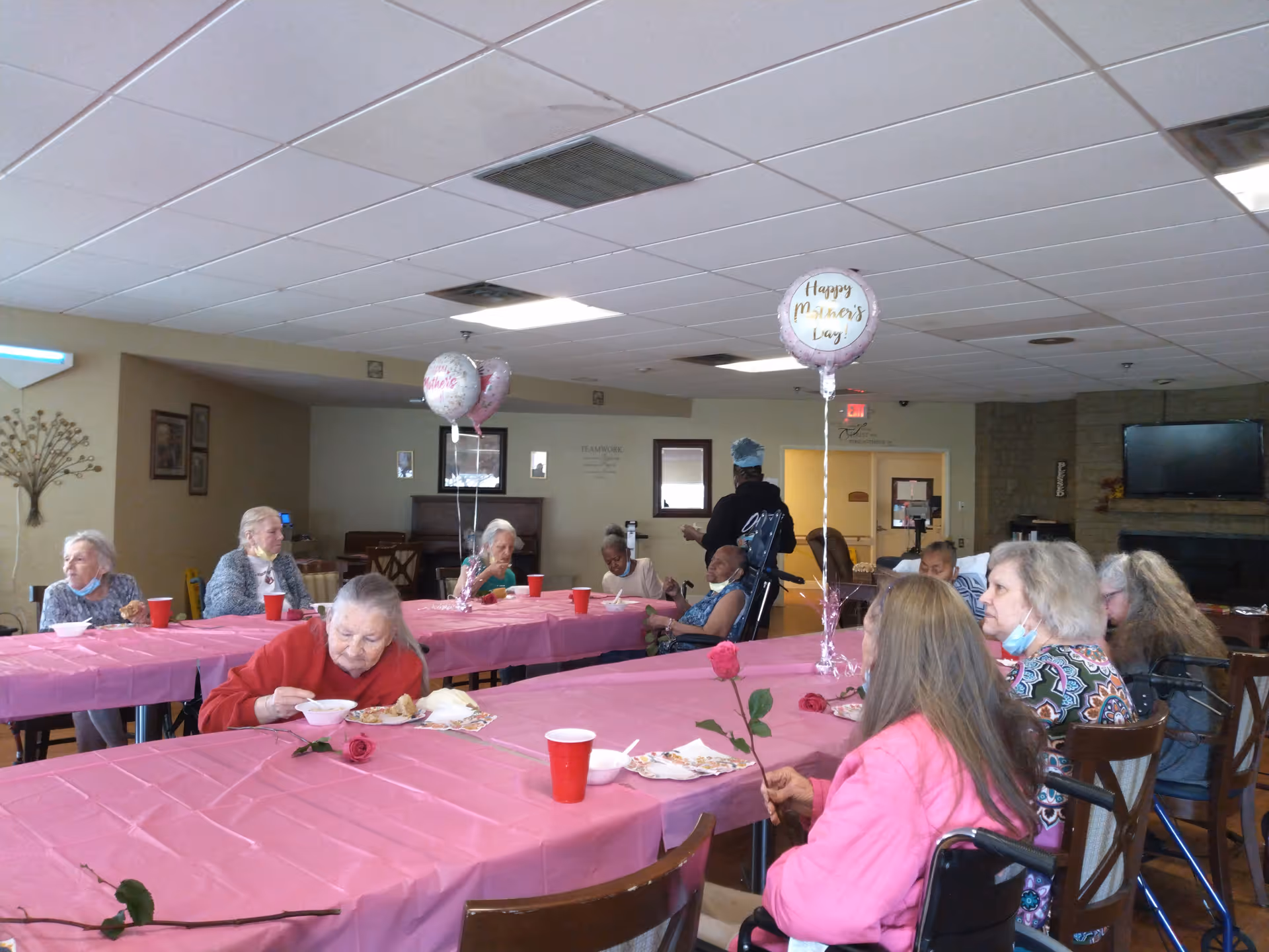 Older adults seated around long tables covered with pink tablecloths, eating and holding roses under 'Happy Mother's Day' balloons in a communal dining room.