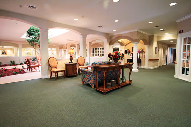 Spacious interior common area with green carpet, beige walls, and white columns. There is a floral-patterned sofa, a wooden table with decorative items, two chairs, and a lamp. The area opens into a courtyard with plants and seating visible through large arches.